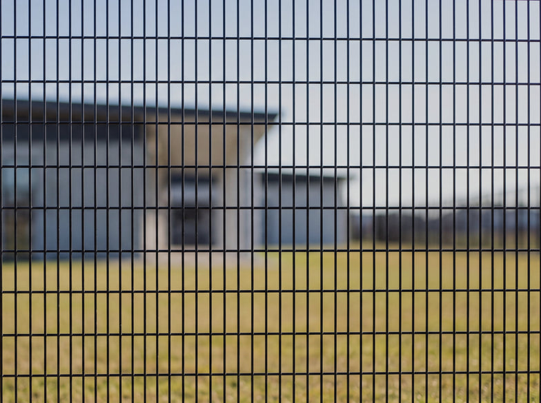 A close-up of a black metal mesh fence gate with a vertical post, set against a blurred outdoor background of a grassy field and modern building.